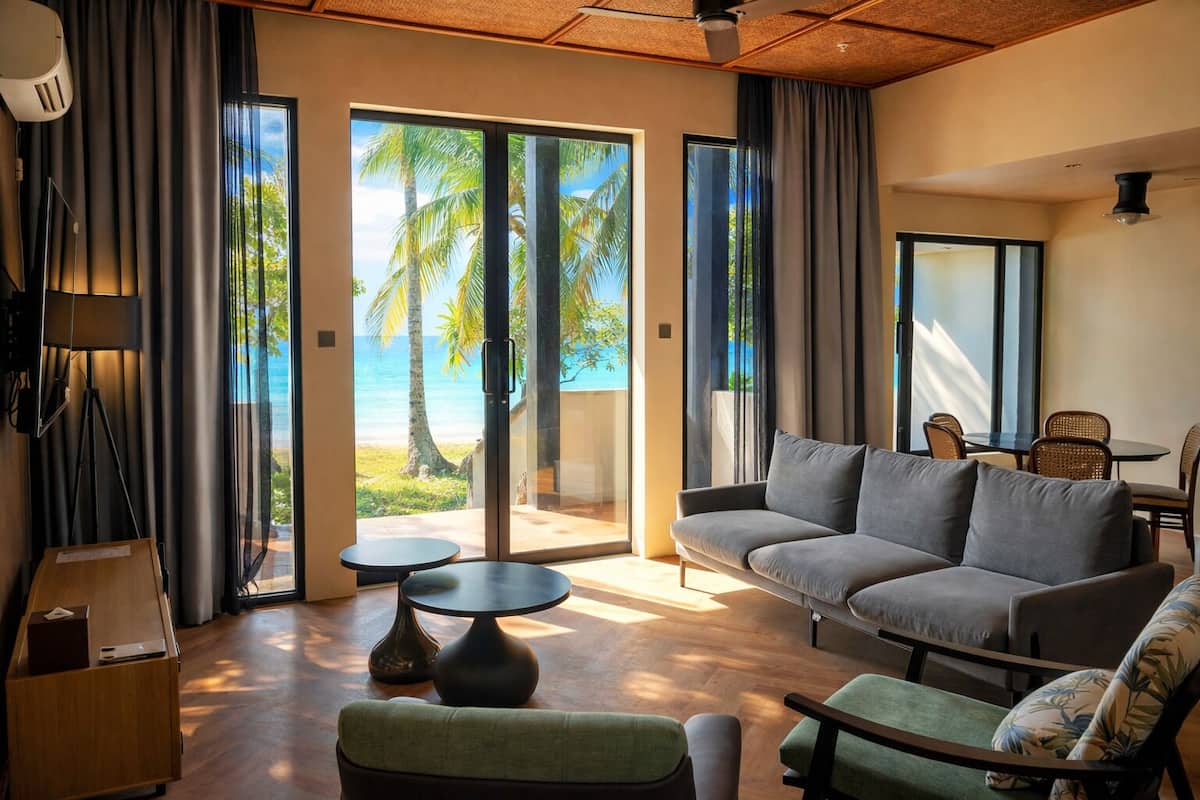 A modern living room with a grey sofa, a small round table, and a green armchair. There is a large sliding glass door leading to a balcony with a view of palm trees and the ocean.