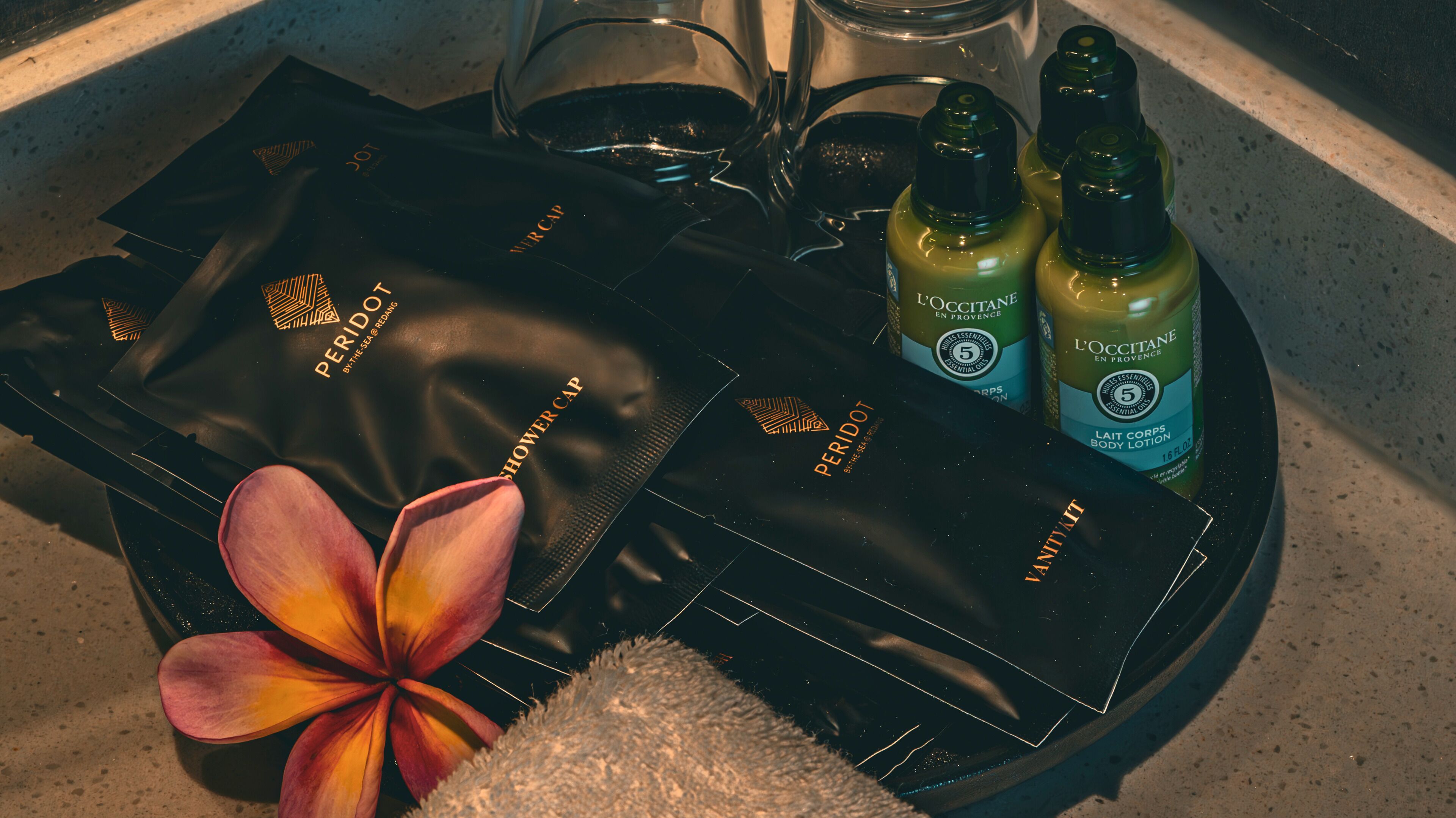A tray with a black leather item labeled "PERIDOT," two bottles of "L'Occitanie en Provence" body lotion, and a tropical flower.