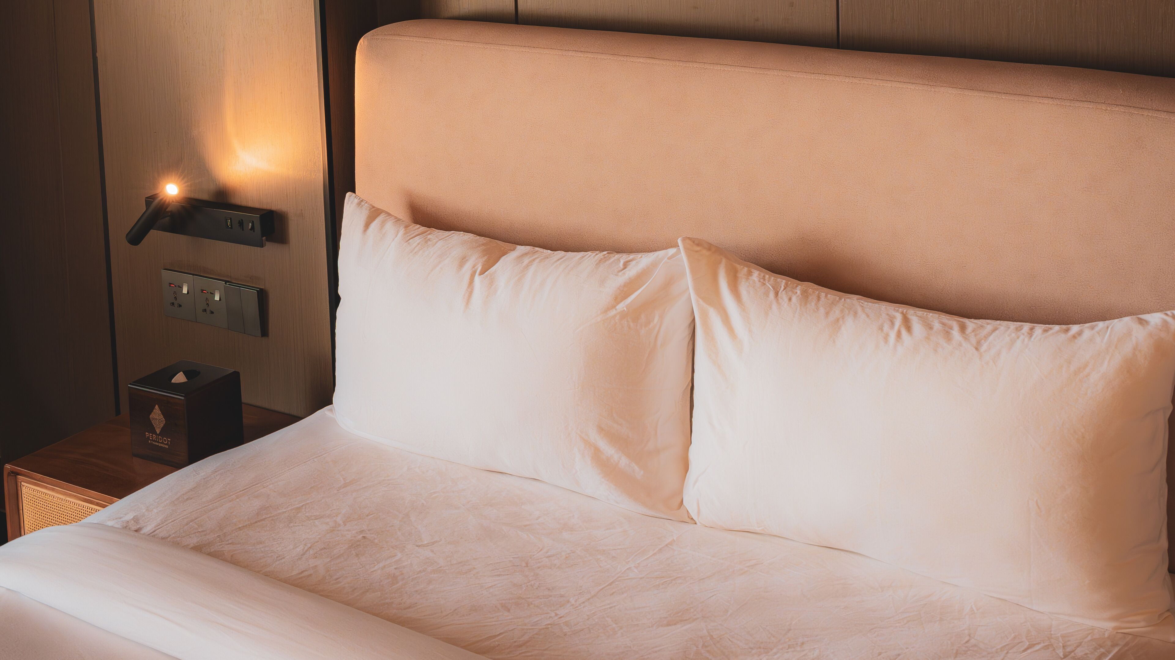 A neatly made bed with a headboard, a bedside table with a tissue box, and a wall-mounted light.
