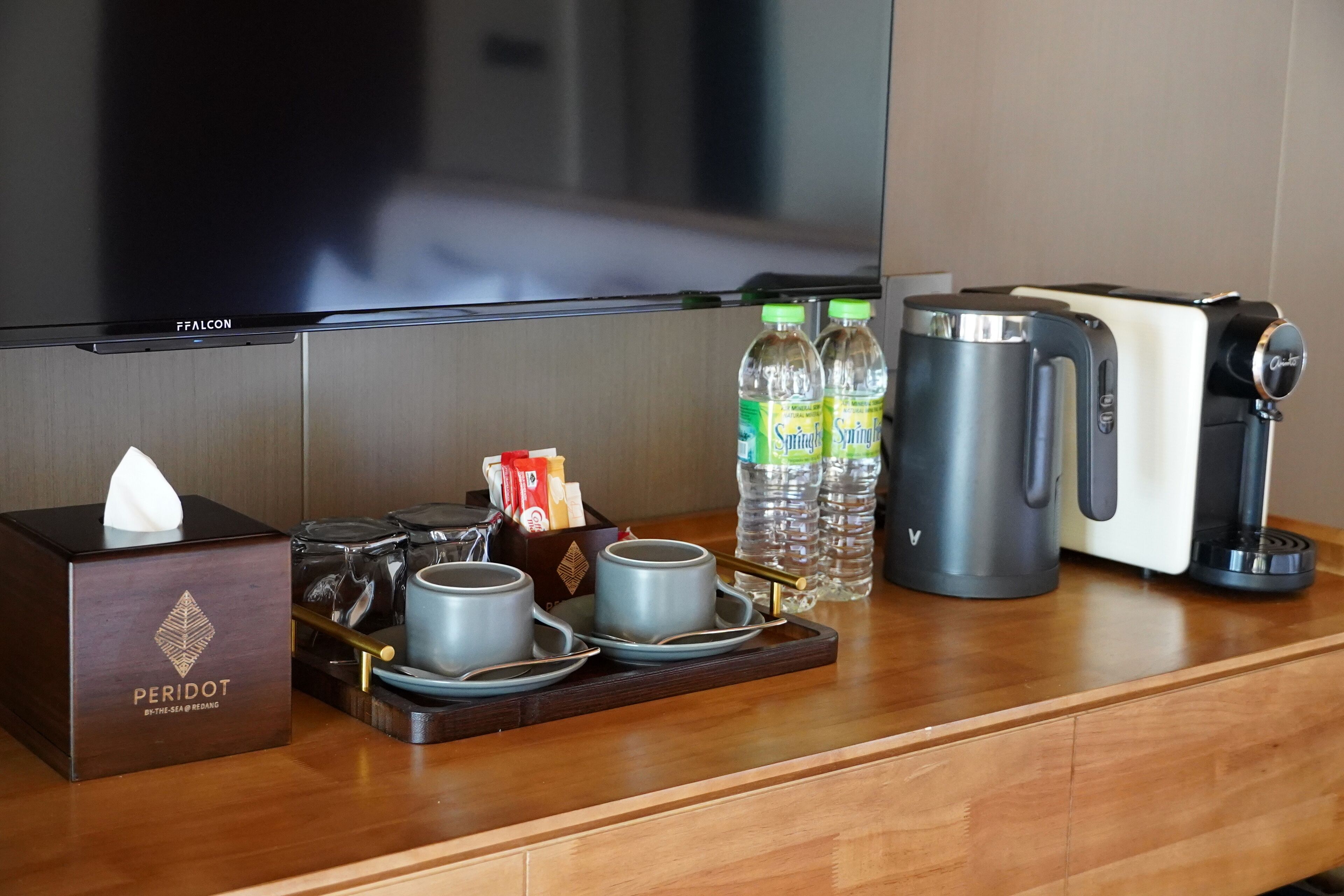Habitación de hotel con minibar que incluye un dispensador de pañuelos, una bandeja con vasos y una termo, agua embotellada y una cafetera.