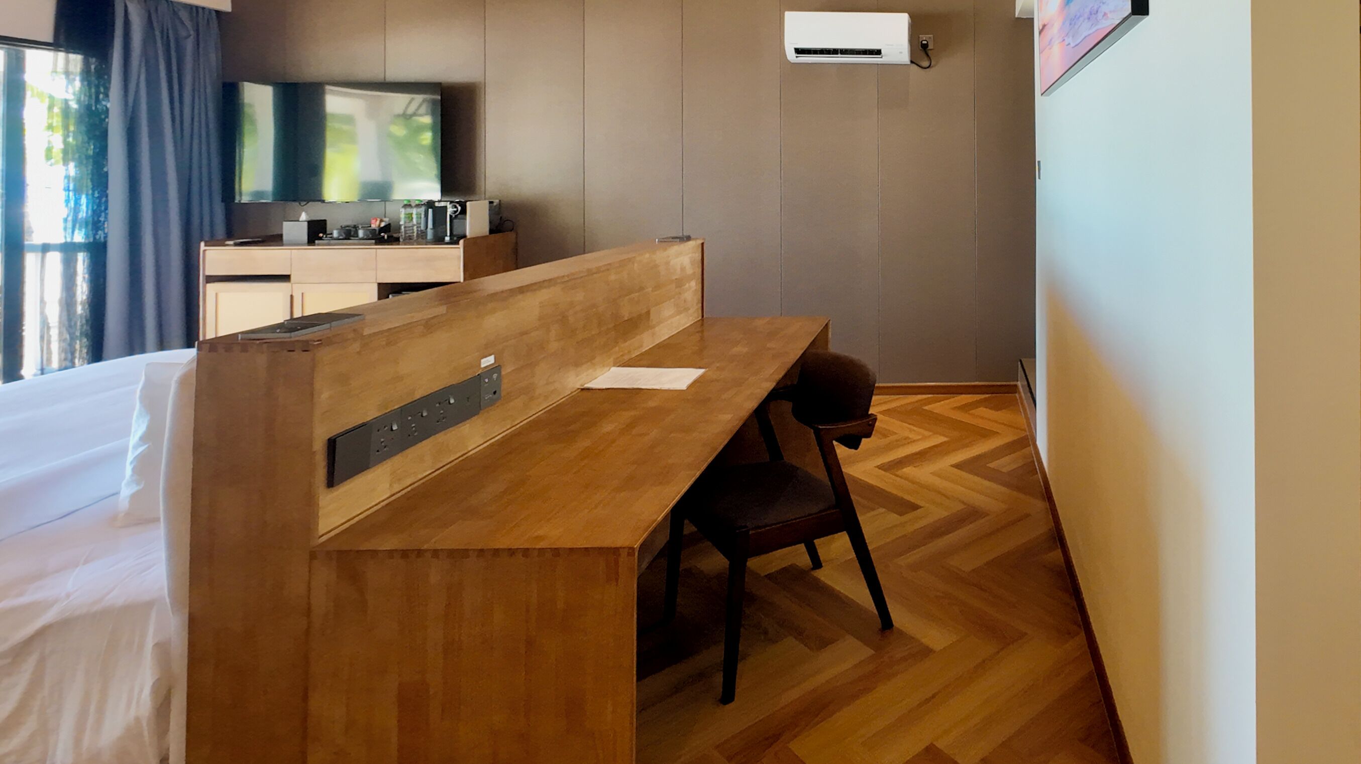 A hotel room with a wooden desk, a chair, a bed, a TV, and an air conditioning unit.