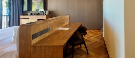 A hotel room with a wooden desk, a chair, a bed, a TV, and an air conditioning unit.