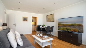A modern living room with a grey sofa, a coffee table, a TV, and a dining area with chairs and a table. - North Sea - Modern and central 2-room apartment with balcony, WLAN. Pets (Sylt)