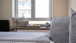 A bedroom with a window, a radiator, a bed with striped bedding, a pillow with a number, and a pitcher on the windowsill. - North Sea - Modern and central 2-room apartment with balcony, WLAN. Pets (Sylt)