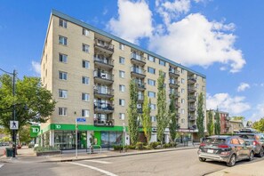 Ein mehrstöckiges Wohnhaus mit einem grünen Laden und einem Fußgängerüberweg, daneben ein geparktes Auto auf der Straße.