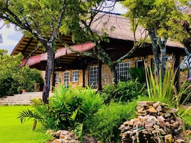 Una casa rústica con techo de paja, paredes de piedra y un jardín con diversas plantas y una formación rocosa.