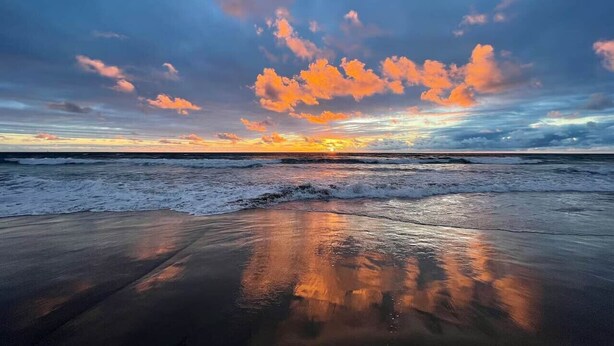 Una playa con olas, un atardecer y un avión en el cielo.