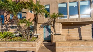 Un edificio moderno con fachada de concreto, ventanas grandes y un pequeño espacio exterior con escalones y palmeras.