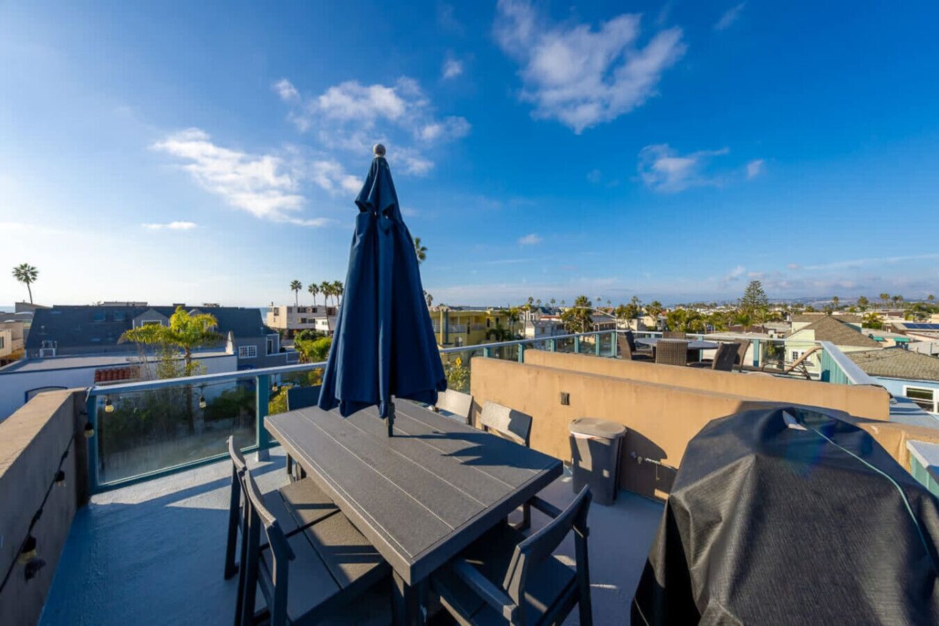 A rooftop terrace with a table and chairs, a covered umbrella, and a view of the surrounding neighborhood.