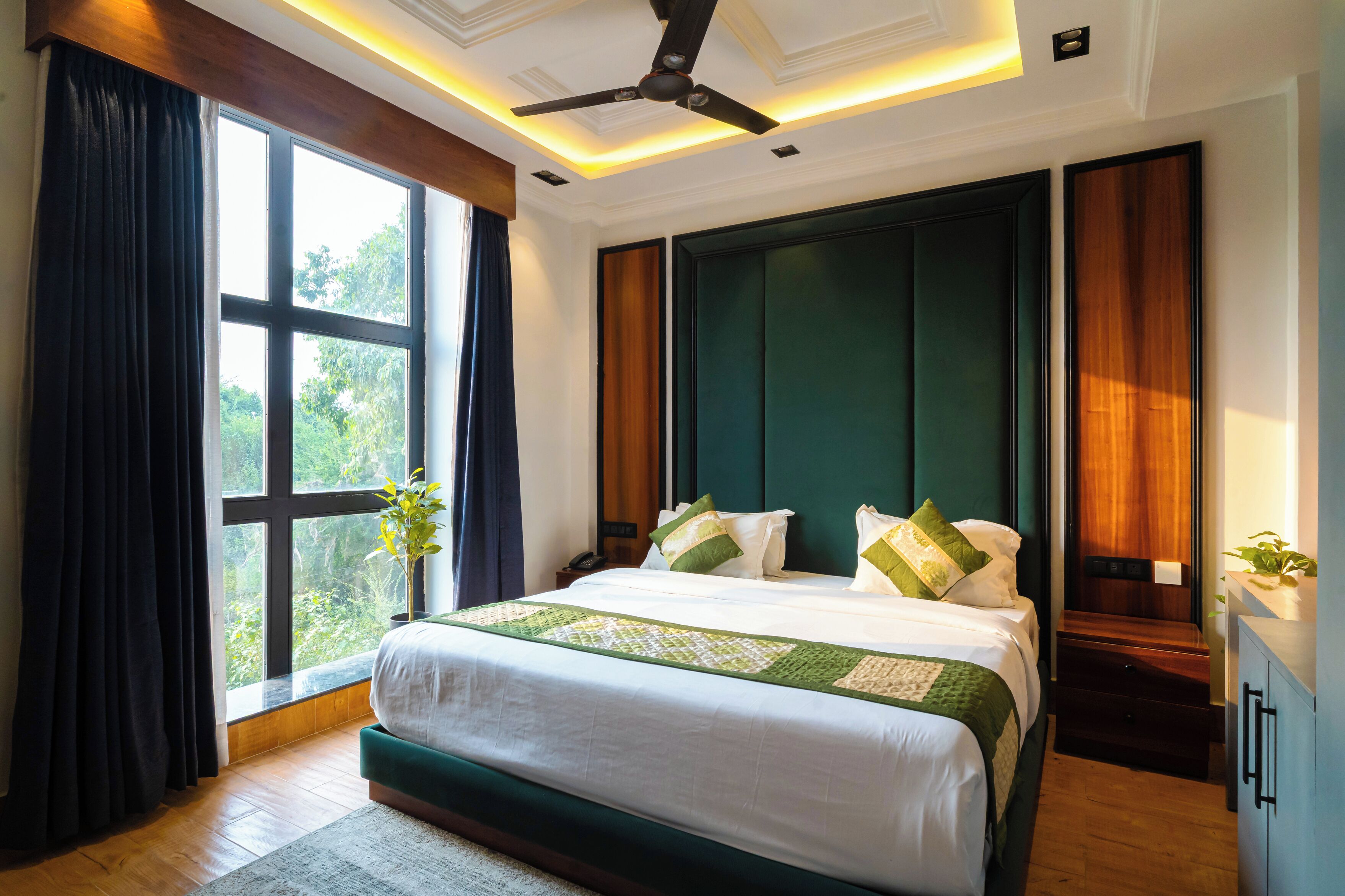 A bedroom with a large bed, a ceiling fan, a window with curtains, and a nightstand with a plant.