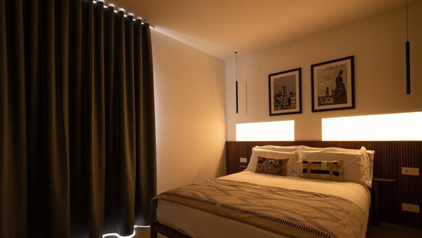 Hotelkamer met een groot bed, donkere gordijnen, een hoofdbord met ingebouwde verlichting en twee ingelijste foto's aan de muur.
