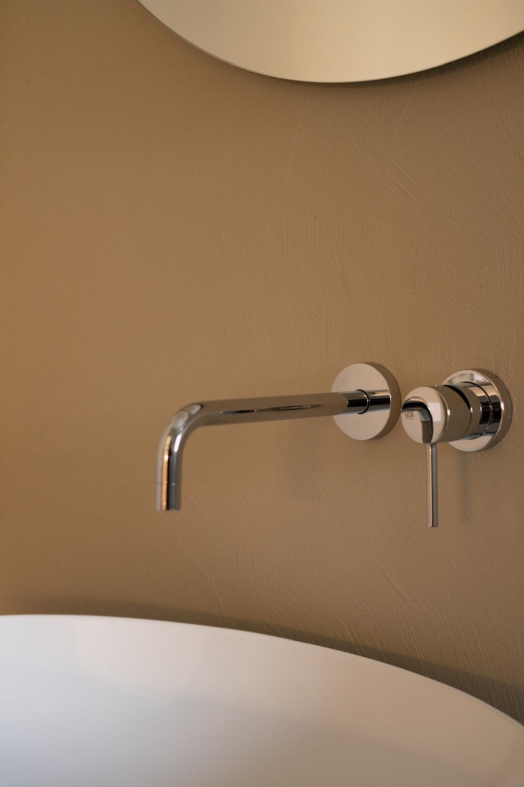 Baño con grifo cromado sobre una pared color beige, un lavabo blanco y un espejo redondo encima.