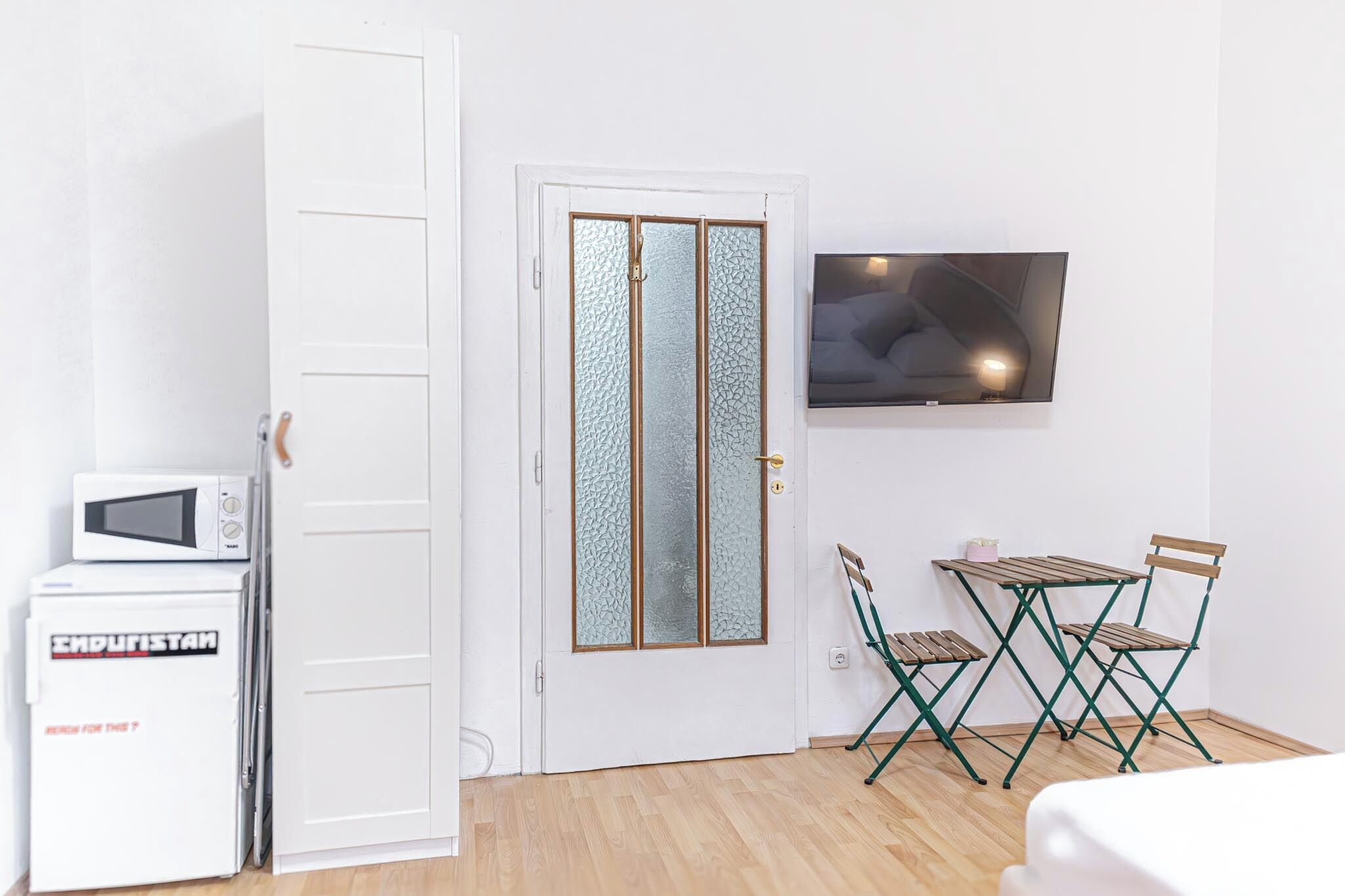 A compact room with a white door, a microwave, a refrigerator, a TV mounted on the wall, and a small table with two chairs.