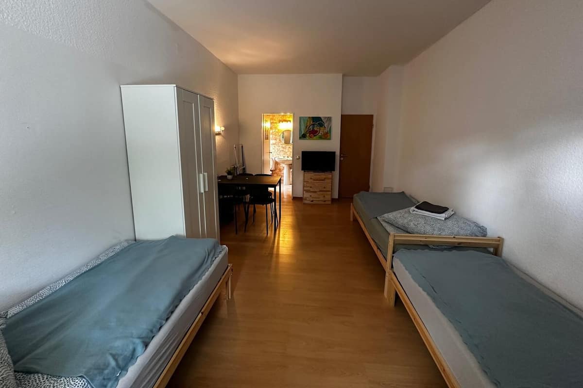 Ein schmales Zimmer mit zwei Einzelbetten, einem Holztisch mit Stuhl, einem kleinen Tisch und einem Fernseher.