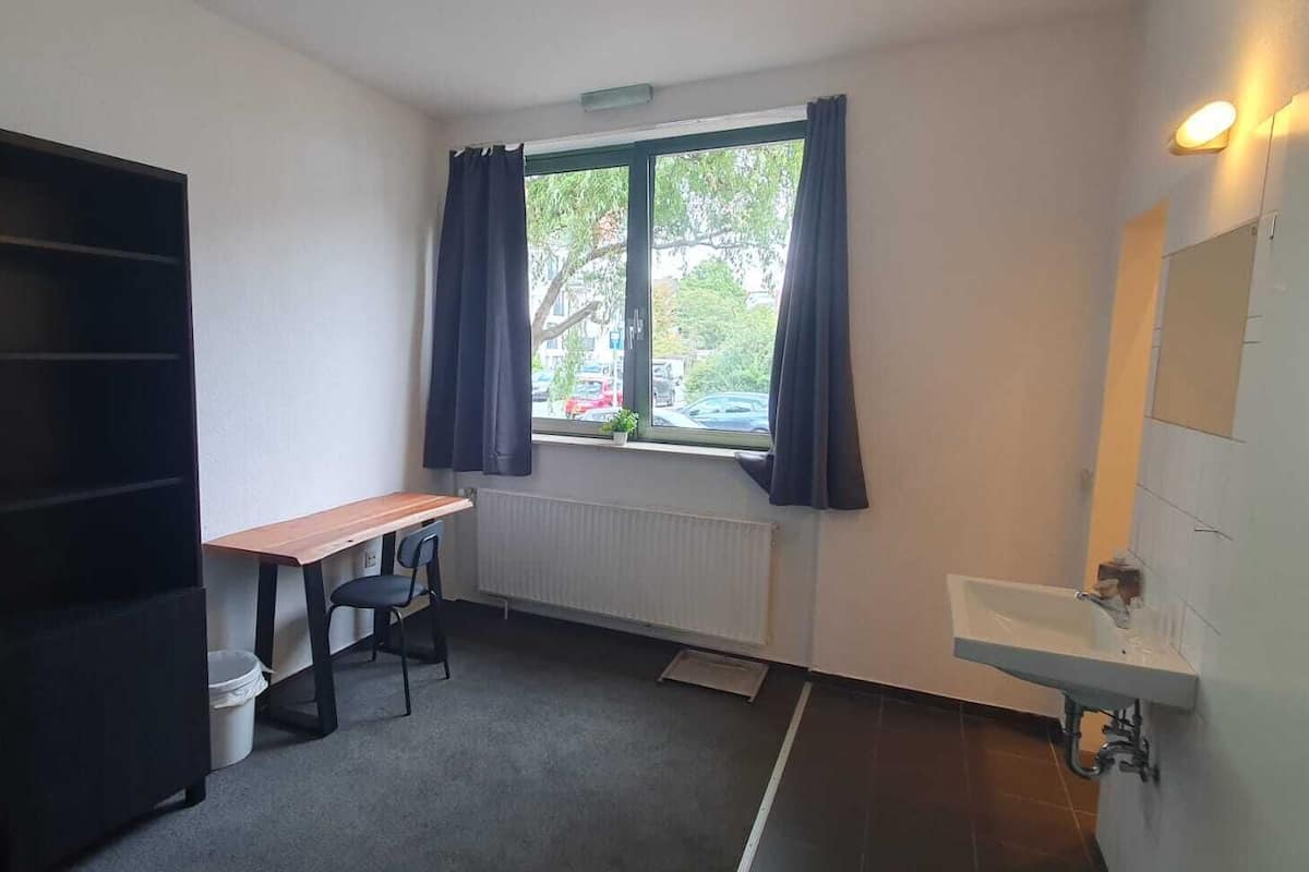 Ein kleiner, einfach eingerichteter Raum mit Fenster, Heizkörper, Schreibtisch und Waschbecken.