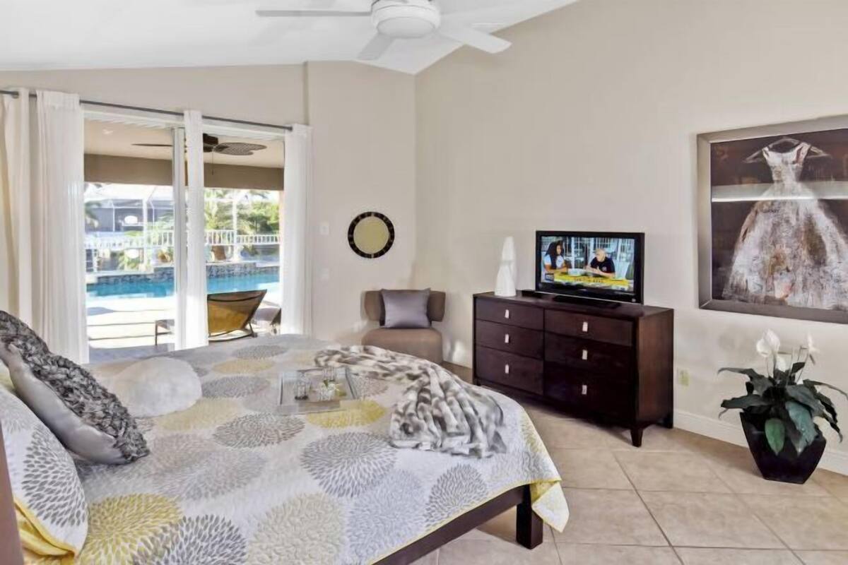 Ein Schlafzimmer mit Bett, Fernseher, Schminktisch, einem Gemälde und Blick auf einen Pool.