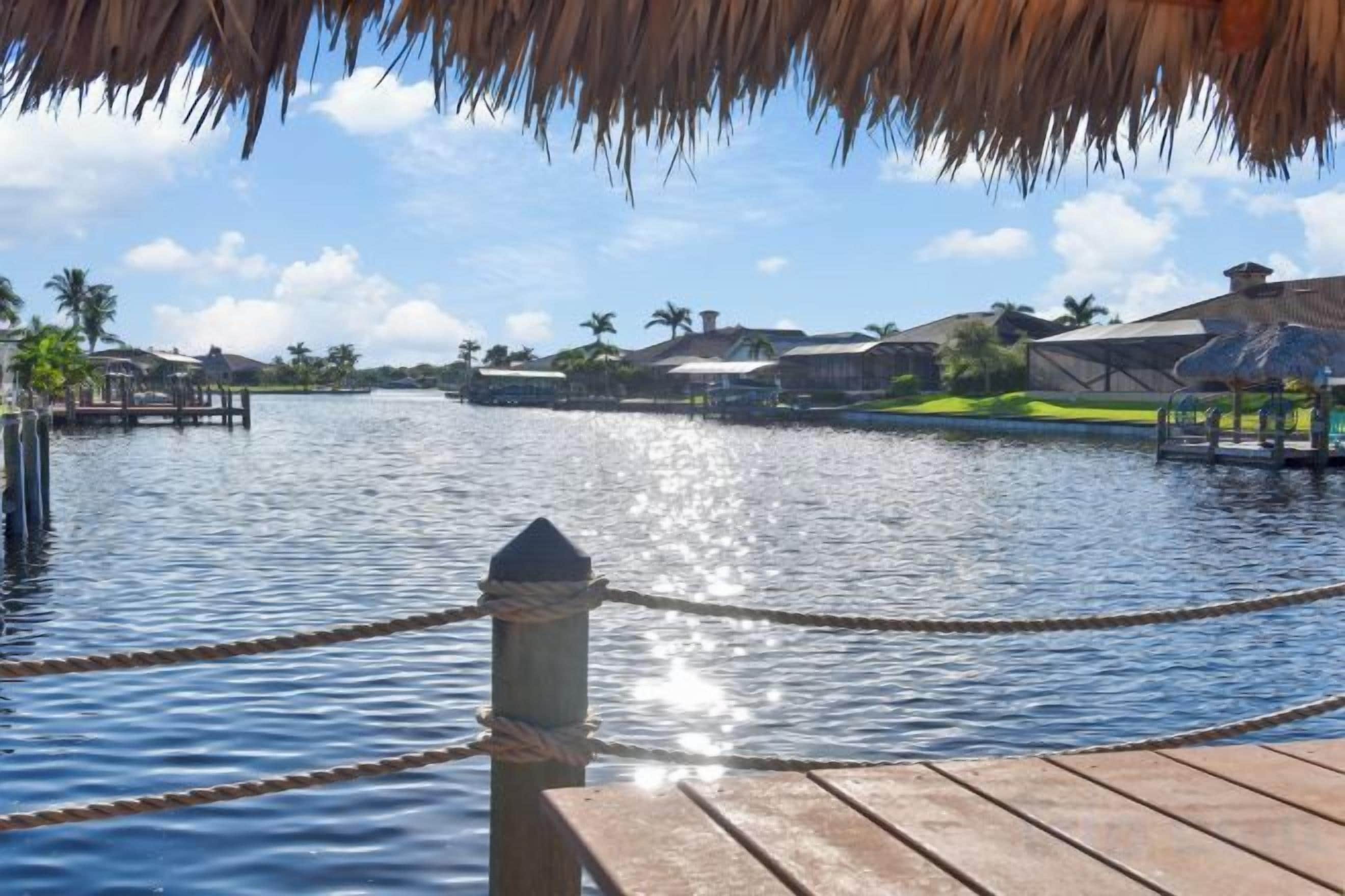 Un muelle de madera con una barandilla de cuerda, un techo de paja y vista al agua con edificios y palmeras al fondo.