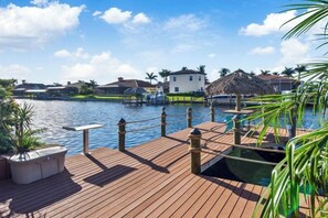 Un muelle de madera con una barandilla de cuerda, una estructura de techo de paja y una masa de agua con edificios al fondo.