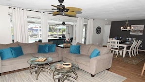 A living room with a beige sofa, two glass coffee tables, a ceiling fan, and a dining area with white chairs and a table. - Coral Sunshine - large south terrace with pool/spa (Cape Coral)