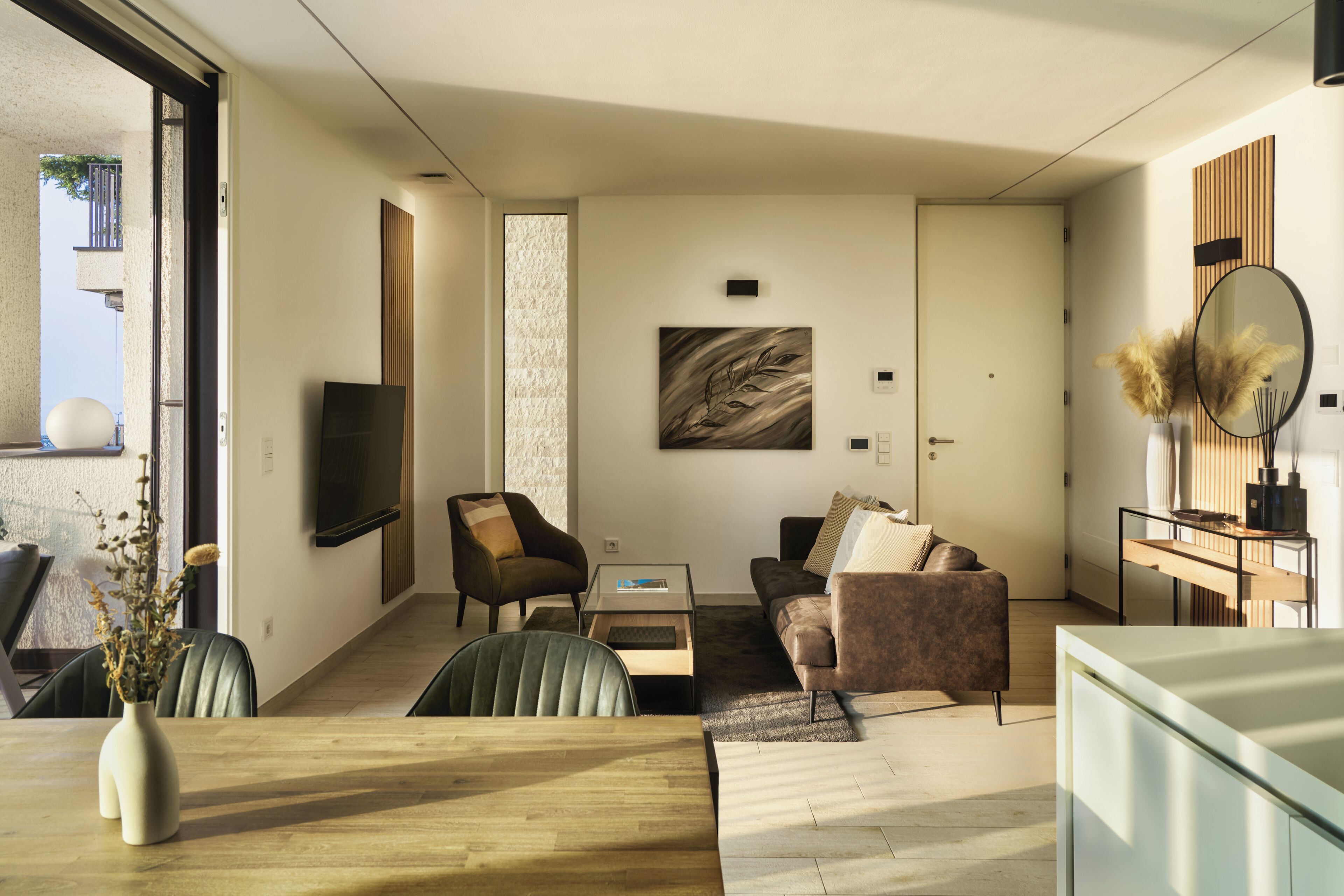 Ein modernes Wohnzimmer mit einer Couch, Sesseln, einem Couchtisch und einem Esstisch. An der Wand ist ein Fernseher angebracht und über der Couch hängt ein Gemälde.