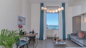 A modern living room with a dining area, a sofa, a coffee table with magazines, a plant, and a view of the sea through large windows. - Alta Marea Apulia sea view (Bari)