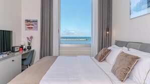 A hotel room with a bed, a desk, a chair, a TV, a window with a view of the sea, and a framed picture on the wall. - Alta Marea Apulia sea view (Bari)
