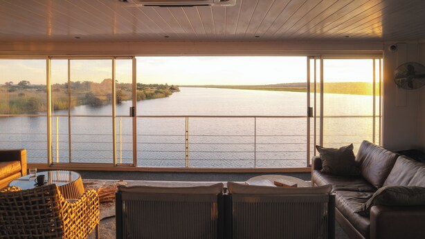 A room with a large sliding glass door leading to a lake view, a leather sofa, a wicker chair, and a small table with a cup. - Zambezi Queen by Mantis  (Kasika)