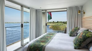 A bedroom with a large window overlooking a body of water and greenery. - Zambezi Queen by Mantis  (Kasika)