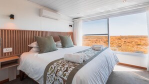 A bedroom with a large bed, a wooden headboard, a bedside table, and a window with a view of a dry landscape. - Chobe Princess By Mantis (Kasika)