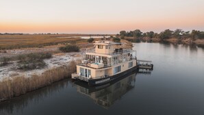 A houseboat named 'CRODE PRINCESS II' docked at a calm river with a flat roof, large windows, and a covered deck area. - Chobe Princess By Mantis (Kasika)