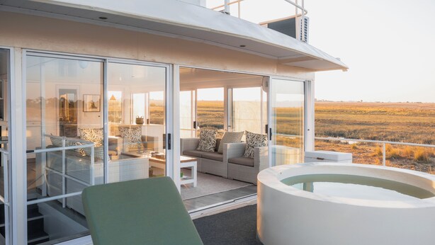 A modern outdoor living area with a glass-enclosed patio, a lounge chair, a hot tub, and a view of a grassy landscape. - Chobe Princess By Mantis (Kasika)