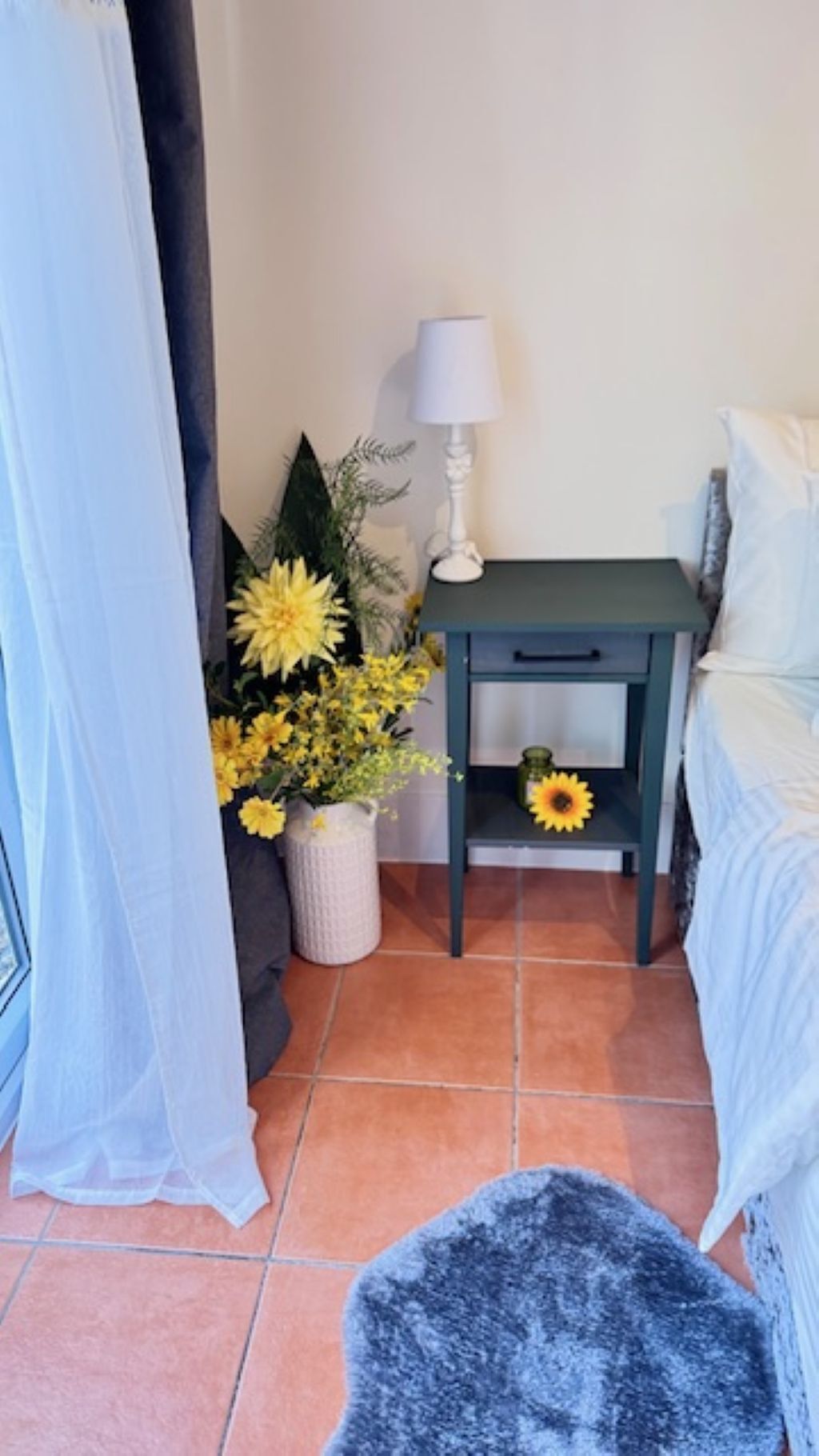 Un dormitorio con cama, una mesita de noche con lámpara, un jarrón con flores amarillas y una alfombra pequeña.