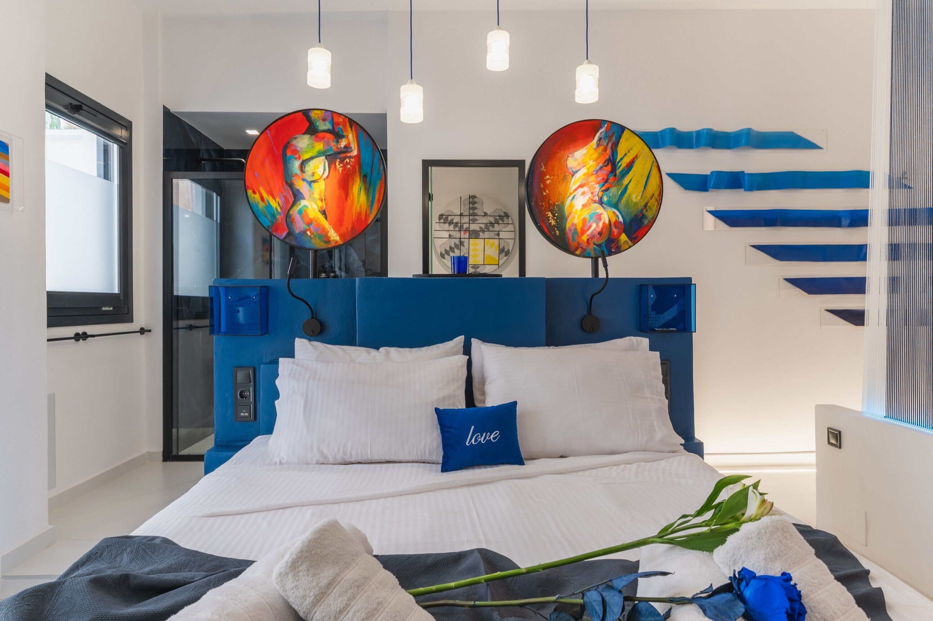A modern bedroom with a blue headboard, white bedding, and two abstract paintings above the bed.