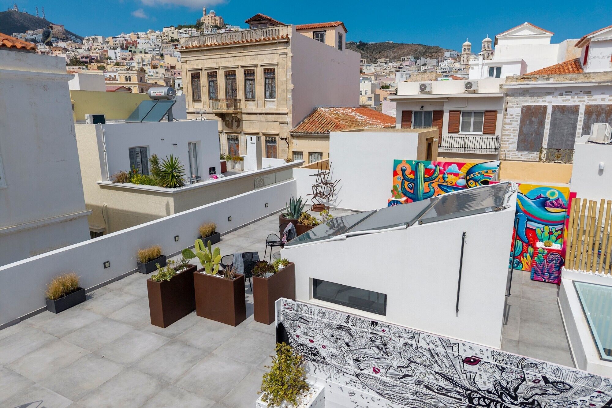 Eine Dachterrasse mit Blumentöpfen, einem großen Wandgemälde und Blick auf die Stadtlandschaft.