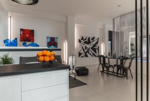 Eine moderne Küche mit schwarzem Arbeitsplatz, weißen Schränken und einer Obstschale. Es gibt eine Essecke mit Tisch und Stühlen, und ein großes abstraktes Gemälde an der Wand.