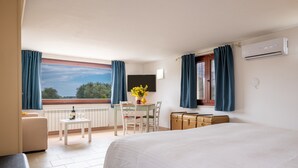 Ett hotellrum med en säng, ett litet bord med en flaska och glas, en stol, en resväska och utsikt över omgivningarna.