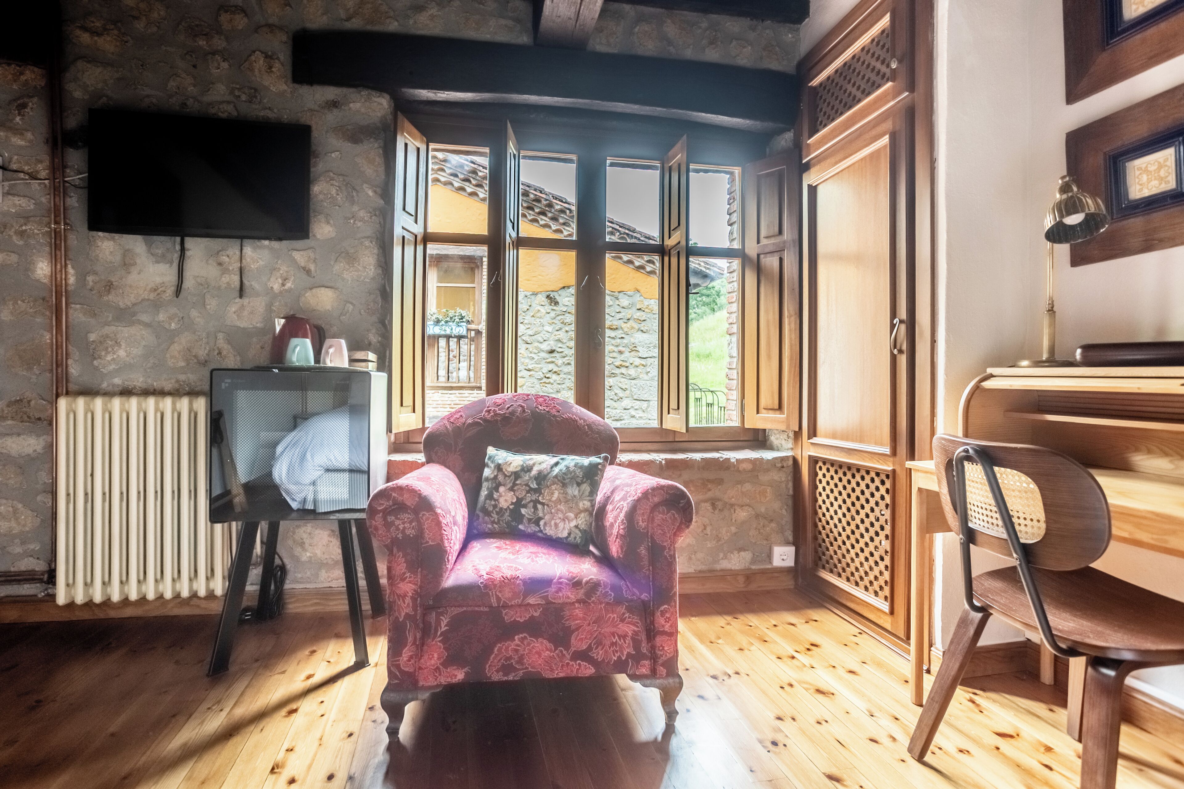 A room with a wooden floor, a red armchair with a cushion, a wooden chair, a radiator, and a window with a view of a stone wall and greenery.