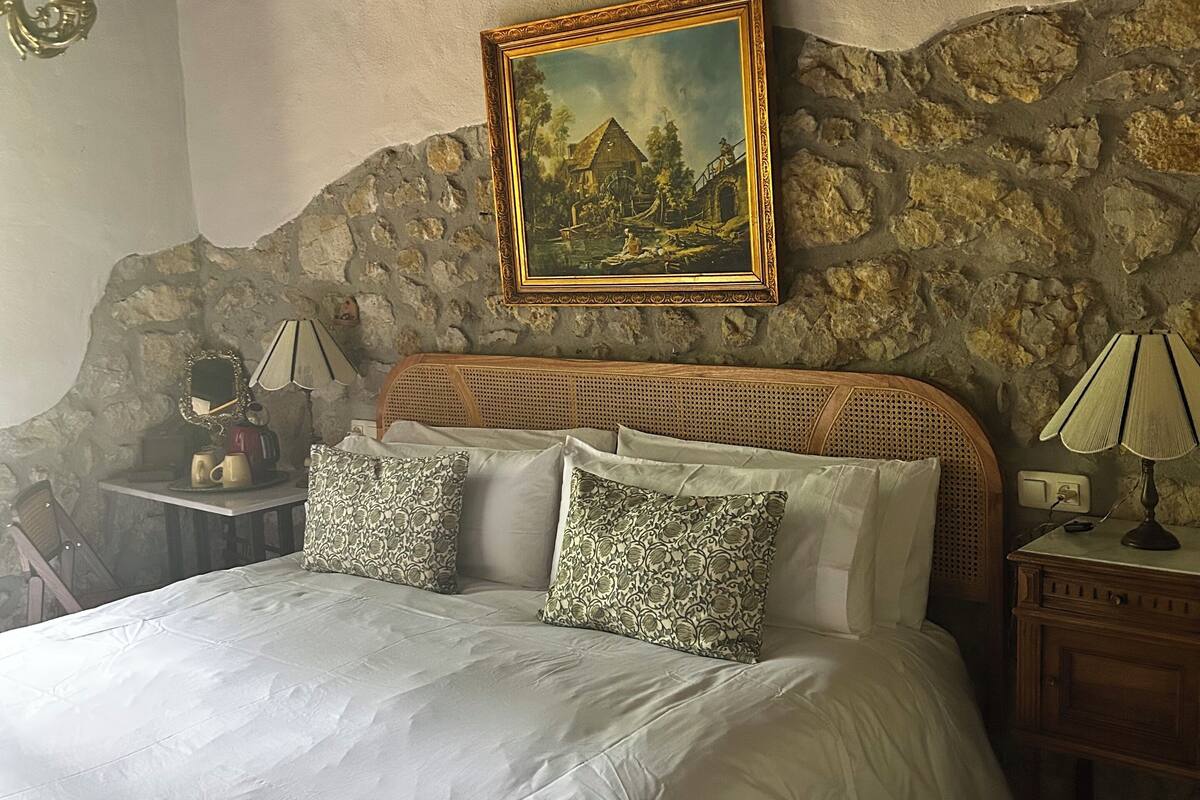 Een bed met witte lakens, een houten hoofdbord en gekleurde kussens. Daarboven hangt een schilderij en op het nachtkastje staat een lamp.