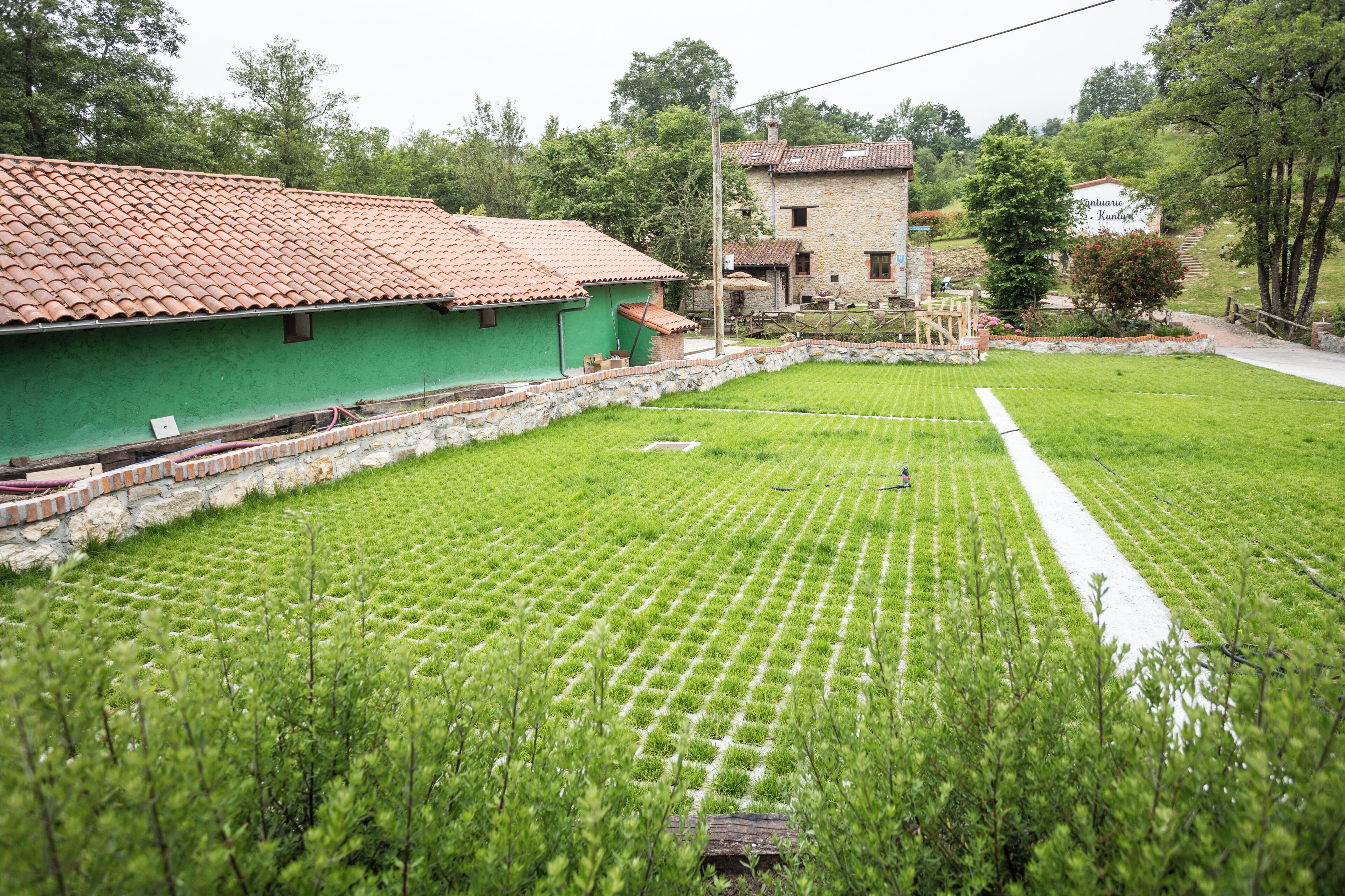 A green building with a tiled roof, a stone wall, and a grassy field with a white path.