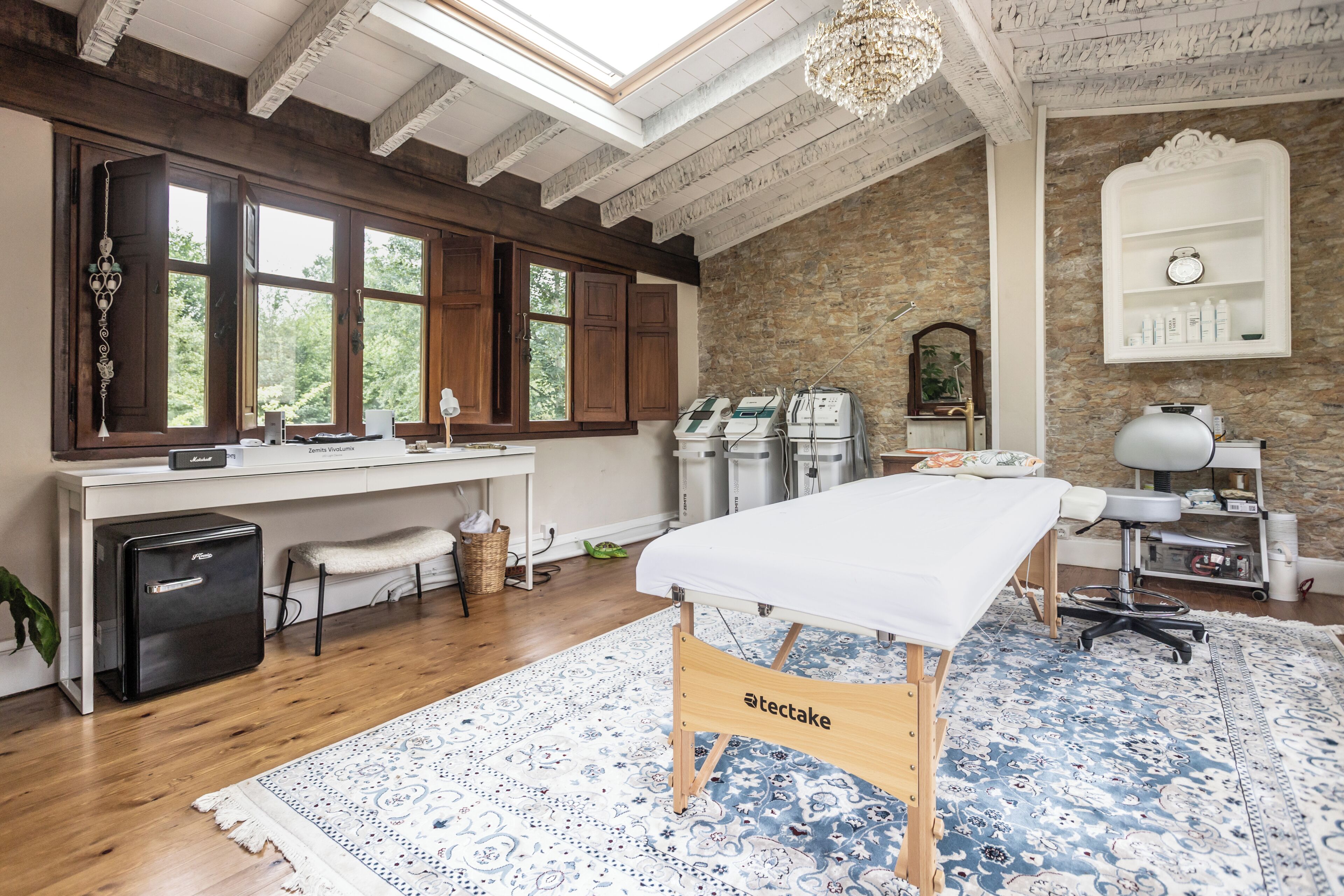 A room with a wooden desk, a massage table, a stone wall, and a large window.