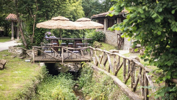 Un pont en bois rustique enjambant un ruisseau, une table à toit de chaume avec des chaises, et un bâtiment en pierre surmonté d’un toit en tuiles.