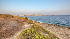 A coastal landscape with a rocky path leading to the sea, clear skies, and distant hills. - Penthouse Paradise in Qawra opposite the beach ! (St. Paul`s Bay)