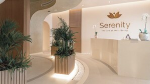 A reception area with a modern design, featuring a reception desk, potted plants, and a wall with the logo and name 'Serenity The Art of Well Being'.