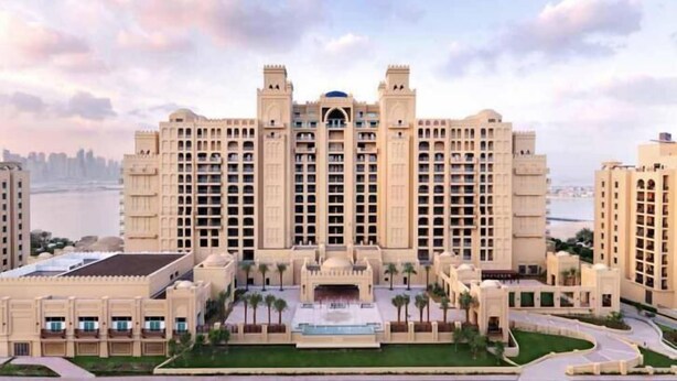 A large, multi-story hotel with a central courtyard, surrounded by palm trees and a waterfront view.