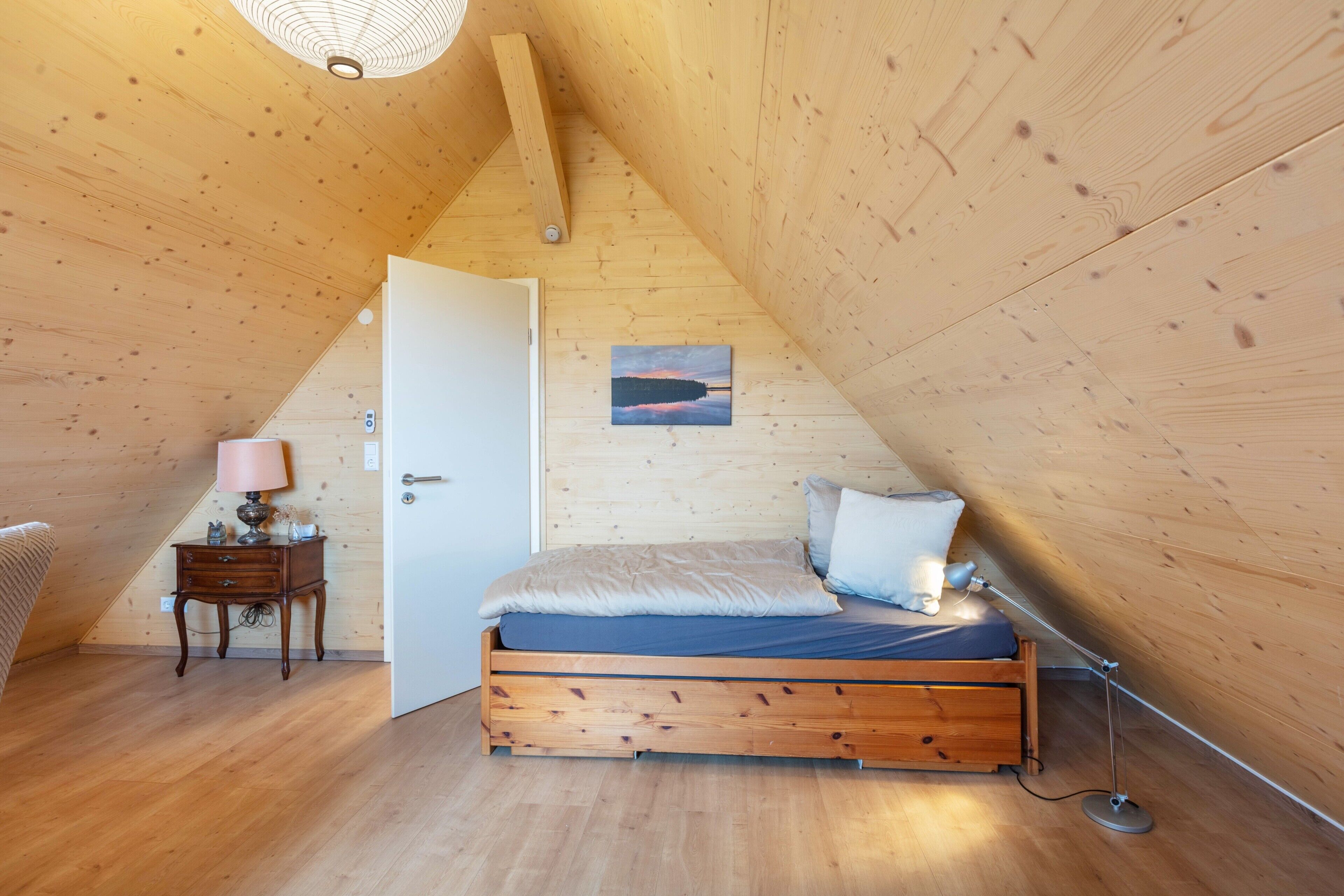 A wooden-paneled attic bedroom with a bed, bedside lamp, and a door leading to another room.