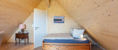 A wooden-paneled attic bedroom with a bed, bedside lamp, and a door leading to another room.