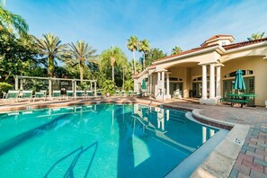 Ein Swimmingpool mit Liegestühlen und Sonnenschirmen, umgeben von Palmen und einem Gebäude mit Säulen.