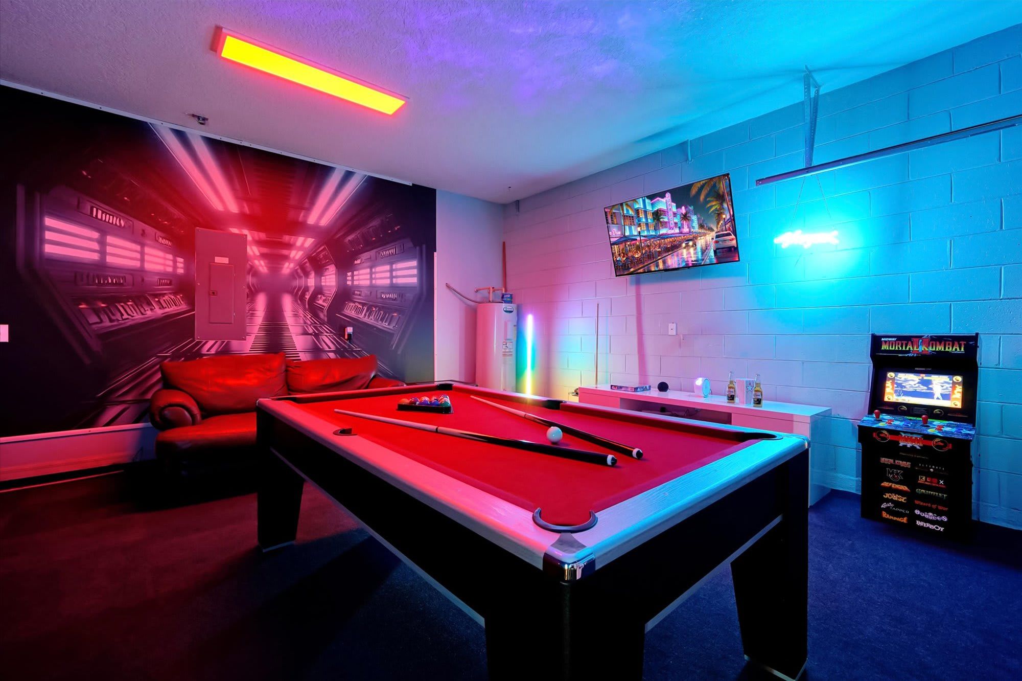Een biljart tafel met een rode viltkleed, een biljartkeu en een poolrek in een kamer met blauwe muren, neonverlichting en een aan de muur bevestigd televisiescherm waarop een spel wordt getoond.