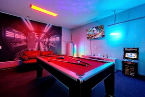A pool table with a red felt surface, a billiards cue, and a pool rack in a room with blue walls, a neon light, and a mounted TV screen displaying a game. - Family-Friendly 6BR Villa w/ Pool, Game Room (Kissimmee)