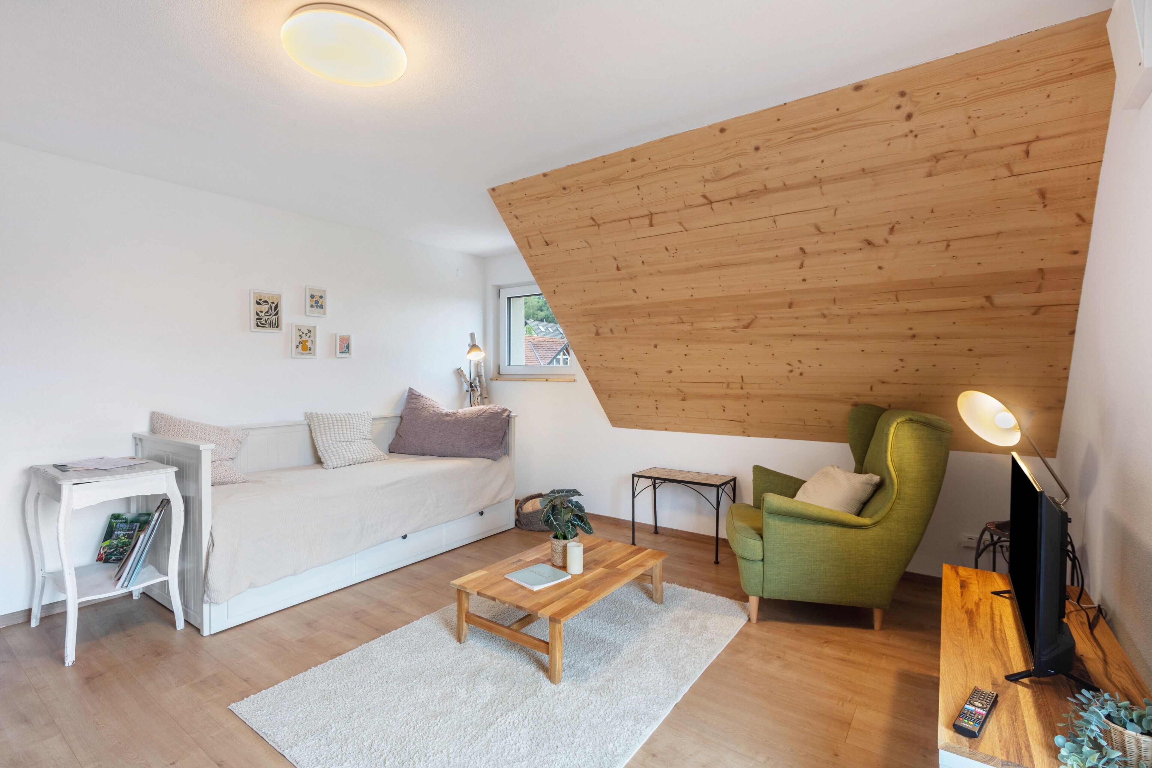 A loft-style room with a sloped ceiling, a sofa, a coffee table, a green armchair, a TV, and a small table with a lamp.